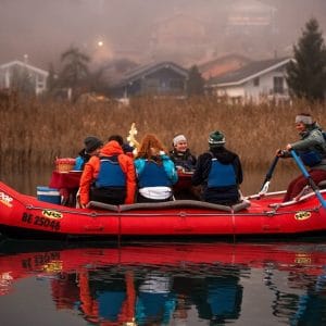 Group of people enjoying raclette on a raft in Switzerland, paddling through a calm river surrounded by misty scenery.